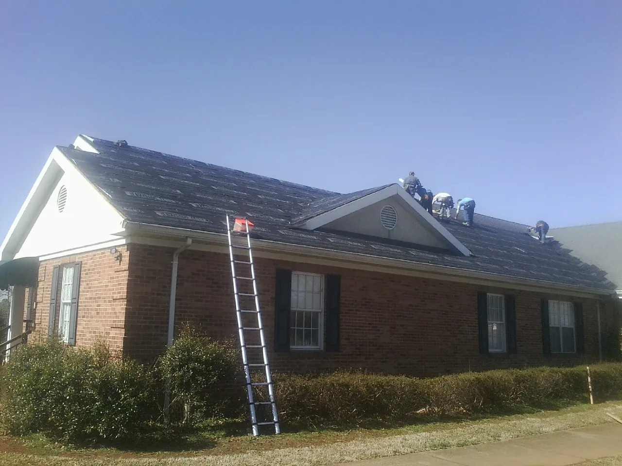 Crew tearing shingles off a brick ranch roof with a ladder set — Emergency Roof Repair work by our crew in East Berlin